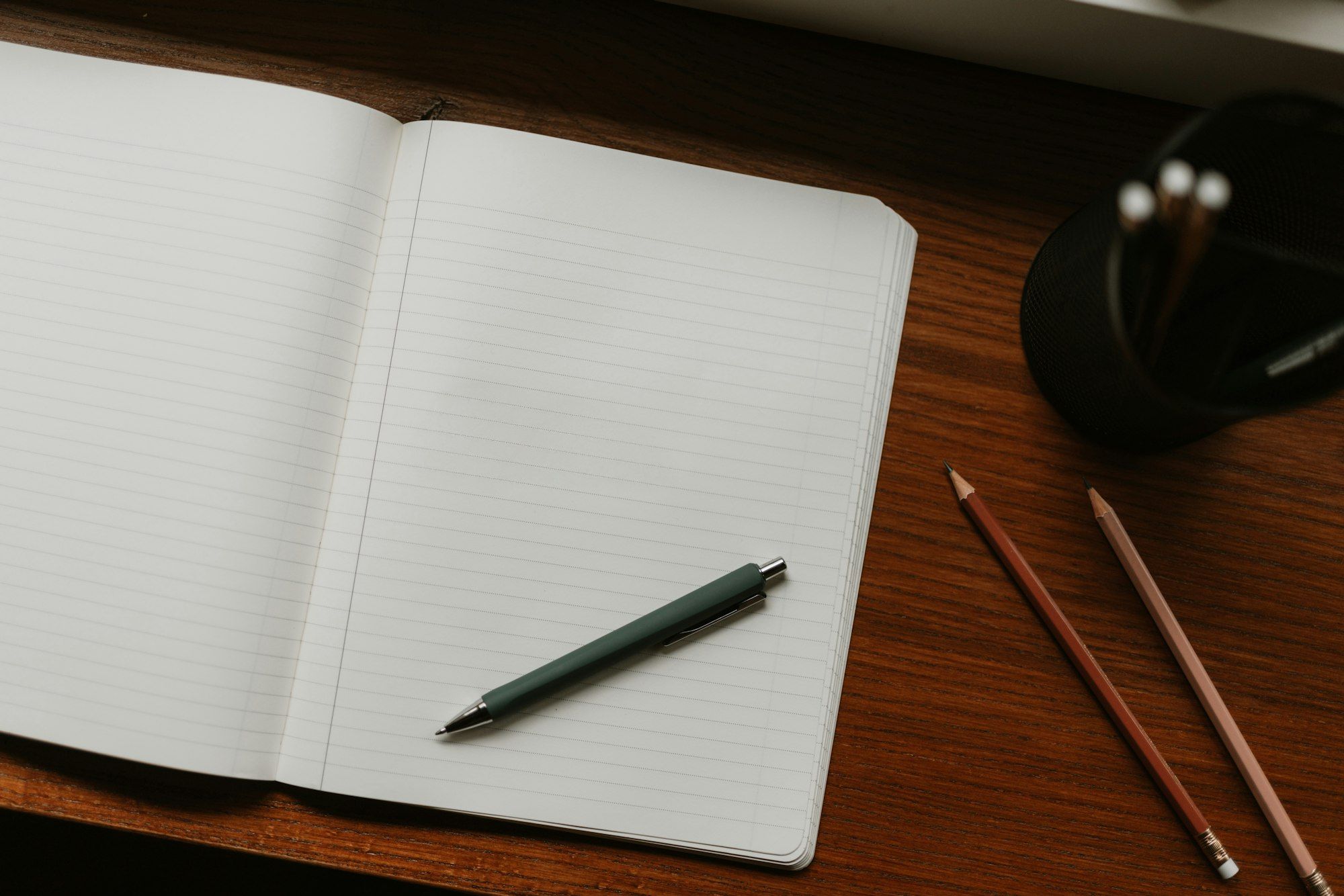 An open notebook with a pen and pencils on a wooden desk.